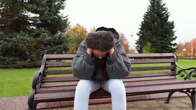 Exhausted young woman sits on city bench in autumn with head lowered in despair. Her body language reflects emotional burnout and overwhelming stress.