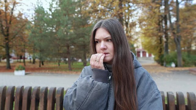Young millennial woman bites her nails on autumn city bench while facing uncertainty about world events. Nervous gestures reflect stress and anxiety.