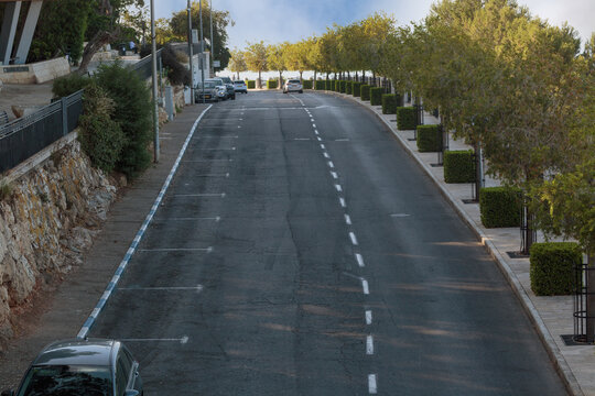 Quiet residential street with empty asphalt road, parked cars, stone walls, sidewalks, and trees in a peaceful urban neighborhood. The elevated view shows road markings and curbside parking areas too
