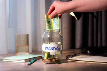Vacation savings in jar. Cash money being added to travel themed savings container. Inspires adventure planning and purposeful financial habits © Krievietka