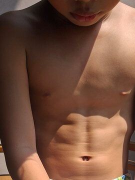 close-up shot of a shirtless boy's torso under natural sunlight, highlighting his stomach and chest with harsh light and shadow casting across his skin.
