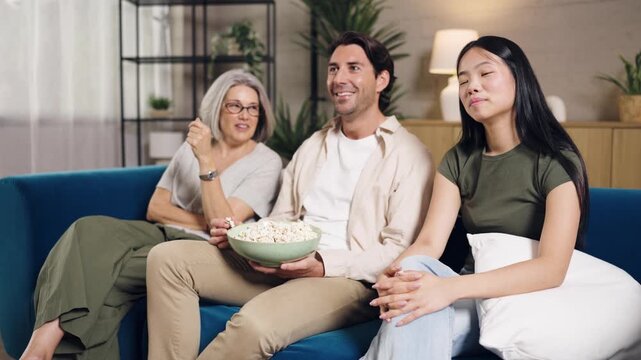 Happy diverse family, including a senior woman, a young couple, watching television and eating popcorn while relaxing on the sofa together