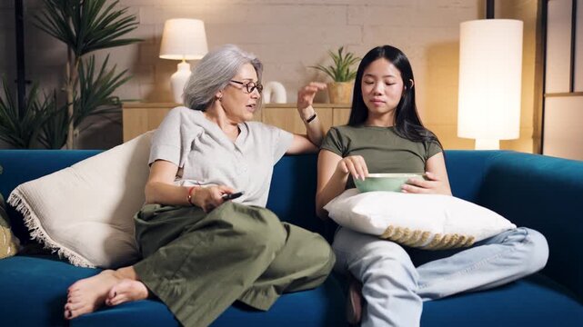Happy diverse family, a senior mother and her young asian daughter, enjoying a movie night, eating popcorn and laughing together on the sofa