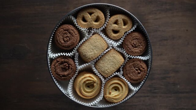 Top view of traditional danish biscuits inside metal can on dark wooden table, rotates. Assorted sweet shortbread pastry cookies in paper cups for christmas holiday marketing