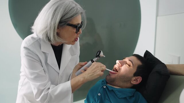 Female physician in a white coat checking a young male patient with a disability. Medical professional using an otoscope during a checkup