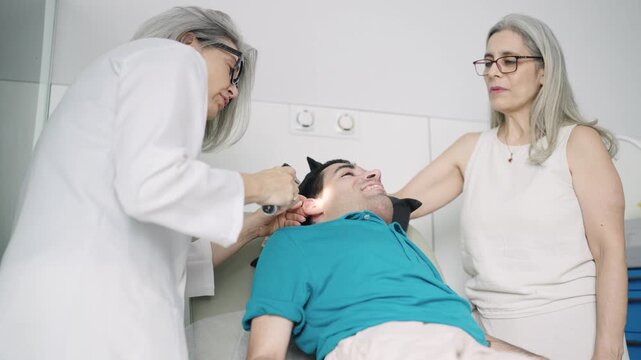 Female audiologist using an otoscope to examine the ear of a young man with a physical disability, accompanied by his mother in a clinic