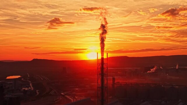 A flare stack sends smoke above an industrial plant at sunset, with the sun low on the horizon and clouds over the distant hills.