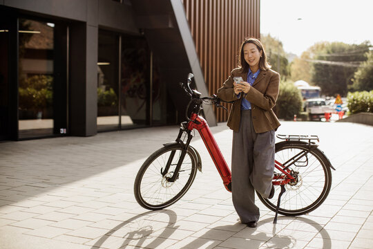 Woman smiles while using phone next to red electric bike in urban setting