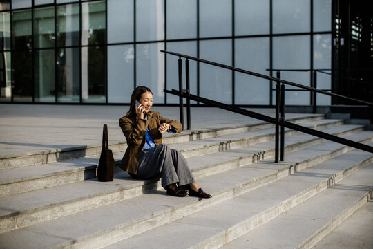 Businesswoman sits on steps and talks on phone while checking her watch