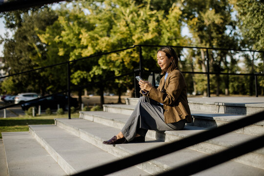Woman sitting on steps using a device in a park with trees and cars in background