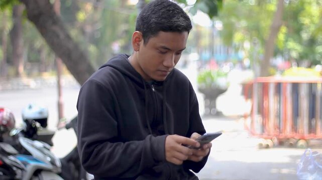 Asian man wearing a black hoodie has a serious expression as he types a message on his smartphone.