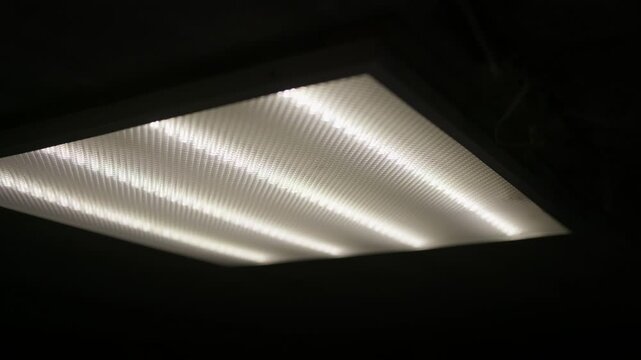 Fluorescent ceiling light glows in a dark room in a low angle view. Static composition captures the bright white illumination shining through the textured diffuser panel.
