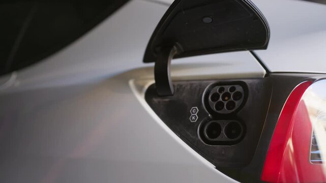 Male hand unplugs charging cable from electric car socket in shot. Selective focus and soft natural daylight highlight the automatic motorized port door closing.