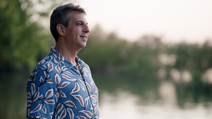 Happy mature man in a patterned shirt smiling while looking out over a calm lake at sunset, enjoying outdoor leisure © DMegias