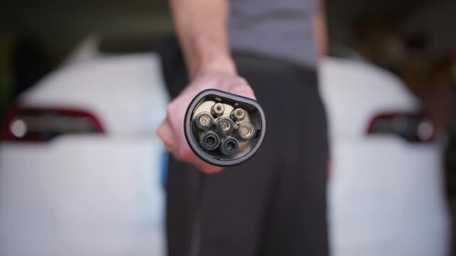 Person holds electric car charging plug in front of vehicle in garage. Selective focus highlights the metallic pins and socket with soft daylight in shot.