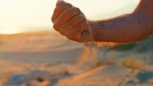 A man pours sand from his palm in slow motion as wind carries the drifting grains. The close shot highlights texture, movement, and nature.