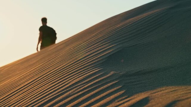 Desert sand dunes reveal a traveler walking in slow motion as wind lifts drifting sand grains and sunlight illuminates the landscape.