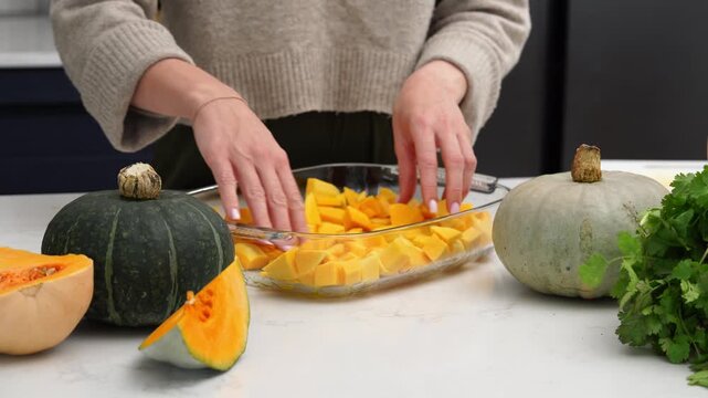 Hands arranging cubed pumpkin in glass baking dish on kitchen counter with whole and sliced squash nearby and fresh herbs visible on side, cozy indoor light, close closeup of preparation scene