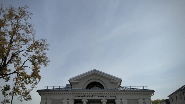 The classical building of the Ukmergė Local Lore Museum featuring prominent white columns, a triangular pediment, an inscribed facade, and dynamic water jets from an active ground fountain 