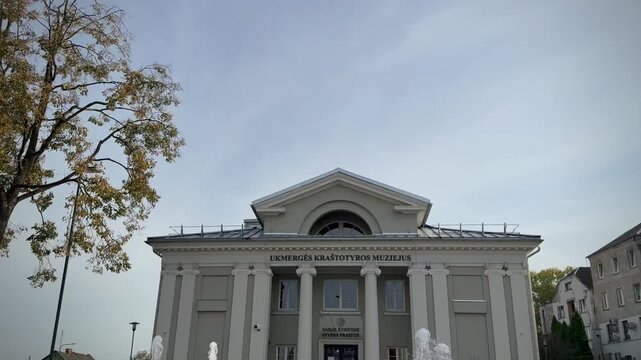The classical building of the Ukmergė Local Lore Museum featuring prominent white columns, a triangular pediment, an inscribed facade, and dynamic water jets from an active ground fountain 