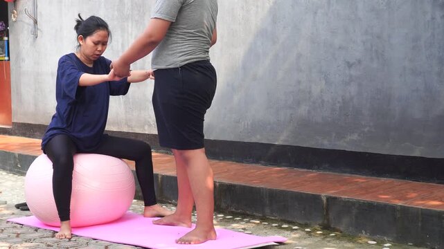 Asian woman exercising on a pink stability ball supported by her partner outdoors.