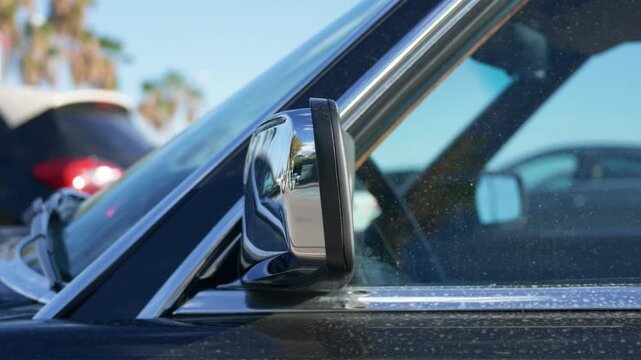 Chrome side mirror of a dark classic car reflecting palm trees, with parked cars blurred behind, French Riviera, France.