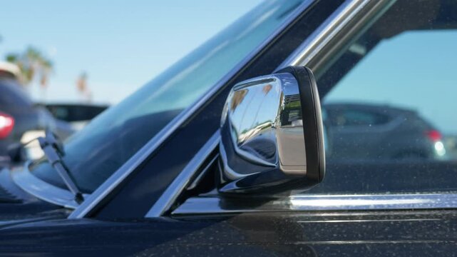 Chrome wing mirror of a vintage black car with palm trees and parked cars in the background on a sunny day, French Riviera, France.