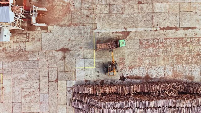 A loader and truck stand beside rows of cut timber on a worksite, seen from above during daytime, with concrete paving and equipment nearby.