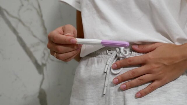 Close-up of woman holding home pregnancy test and touching stomach in bathroom, pregnancy and family planning concept