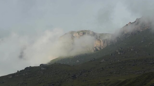 Low clouds drift across a rugged mountain ridge, revealing exposed cliffs and rocky green slopes beneath a shifting overcast sky