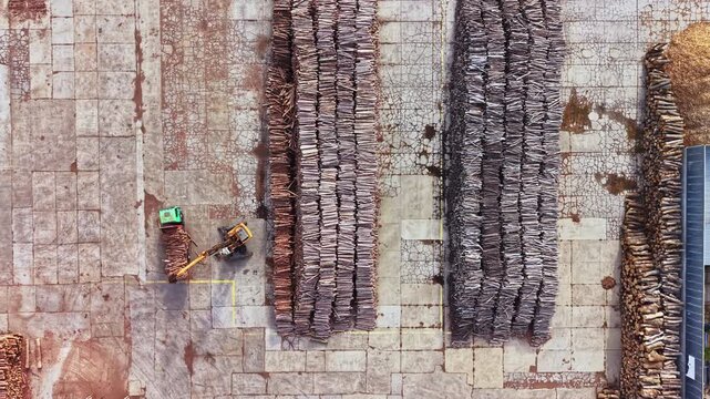 Two workers handle bundled branches on a paved work area beside long rows of stacked materials, seen from above during daytime.