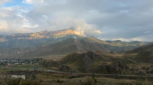 Sweeping mountain valley landscape with rolling grassy hills, winding road, forested slopes, distant village, and cloud covered peaks in a scenic highland panorama