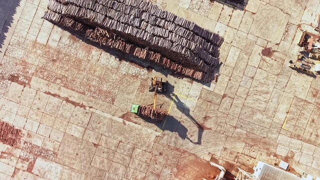 A worker carries materials across a stone paved site beside stacked roof tiles, with long shadows crossing the ground during daylight.