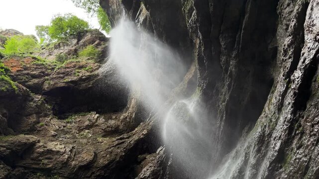 Water cascades down layered rock covered with moss as sunlight filters through a shaded gorge, highlighting the tranquil natural landscape