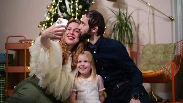 A mother, father and their young daughter sit together on the floor beside a decorated Christmas tree and take a selfie with a smartphone. 