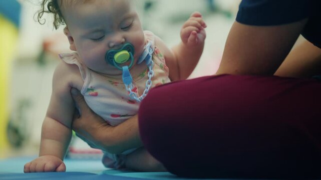 Baby girl with a pacifier pushing up on one arm while a kneeling therapist supports her body on a blue mat in a therapy room, French Riviera, France.