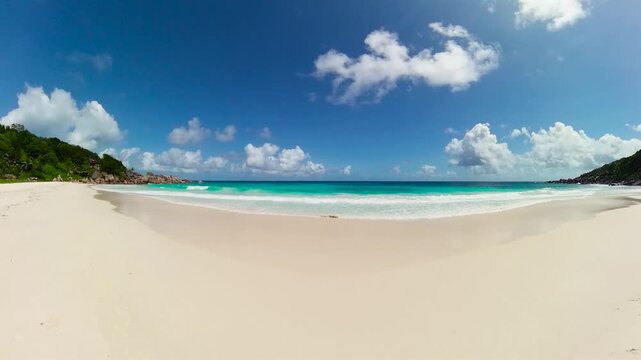 White sand beach with turquoise ocean waves under a bright blue sky with scattered clouds. Seychelles, La Digue. Petite Anse.