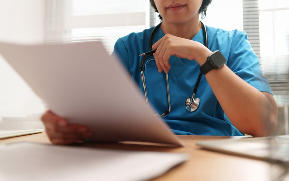 Healthcare professional in blue scrubs with a stethoscope reviews printed medical documents, Clinical setting suggests patient record review and daily care planning.