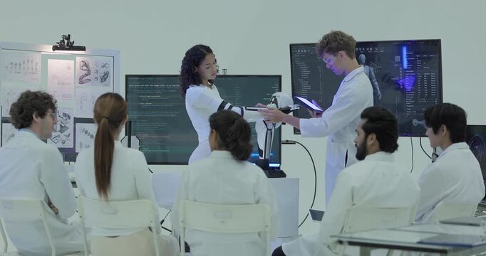 A woman wearing the robotic arm passes the forearm module to an engineer with a tablet, presenting it to the seated team as the humanoid robot specification stays on the screen.