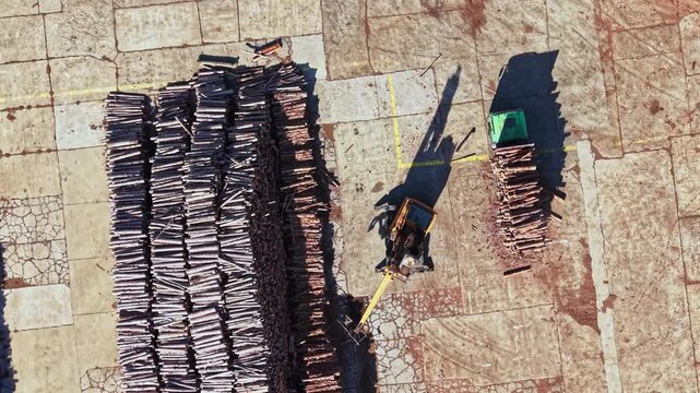Bundles of timber lie in rows on a paved yard as a small loading vehicle moves between the stacks in daylight.