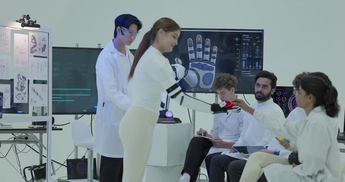 A woman wearing the robotic arm sleeve takes a small block from a seated colleague to test gripping objects of different shapes, with the tactile hand map shown on the screen.