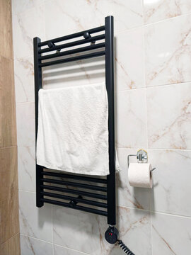 White terry towel hanging on black heated rail against marble effect tile wall. Depicts energy efficient home comfort, post-shower drying, bathroom textile care, and modern interior sanitary design.