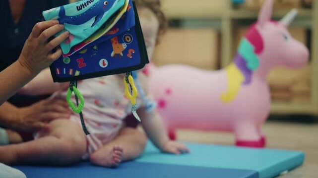 Hand dangling a soft whale cloth book with teething rings in front of a baby girl sitting on a blue mat with a therapist, French Riviera, France.