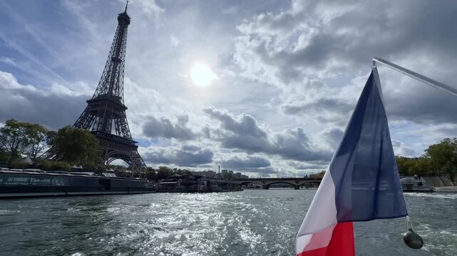 PARIS - Oct 13, 2026 - Wide view of the Eiffel Tower from a sightseeing boat flying the French flag on the Seine River in Paris, France, with dramatic clouds and sunlight reflecting on the water. 1548