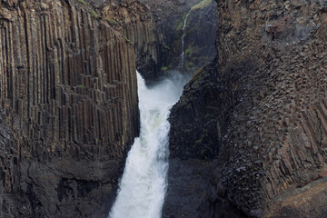 Litlanesfoss cascades through a narrow gorge in East Iceland, where tall basalt columns and fractured cliffs surround white water, drifting spray and small patches of moss. © Artem