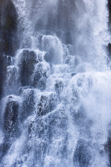 Water tumbles over dark basalt columns at Studlafoss in East Iceland. Fine spray and white cascades fill the vertical frame with an intricate natural background texture. © Artem
