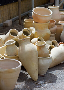Pile of unglazed clay pots and jars in various shapes and sizes stacked outdoors on the ground. Beige terracotta pottery with rustic textures, some pieces broken, in natural daylight. Amphora pots