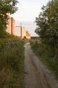 An unpaved dirt trail running through autumn vegetation toward multi-story residential apartment blocks in a provincial town at sunset.