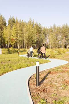 Two mothers walk with baby strollers along a winding path in a sunny autumn park. Back view of women enjoying outdoor leisure with infants.