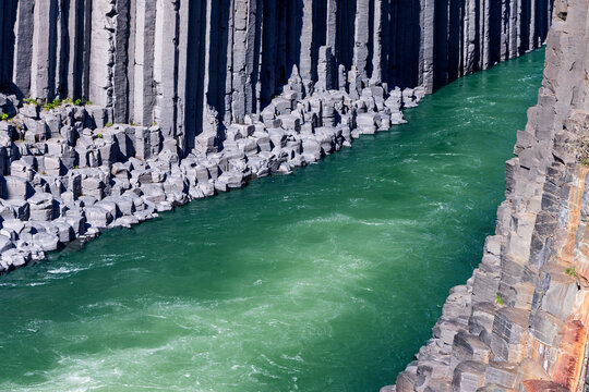 Turquoise water swirls between tall basalt columns in Studlagil canyon, East Iceland. The Jokla river cuts through fractured volcanic rock with orange staining on the near wall.